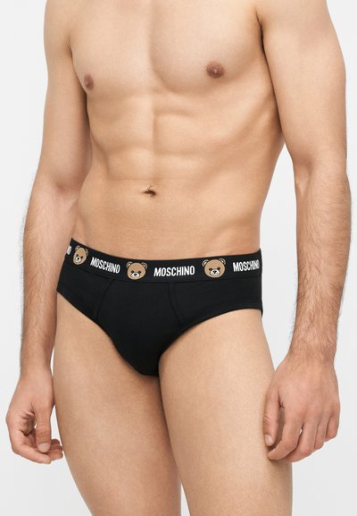 Γυμνασμένος ανδρικός κορμός που φοράει μαύρα εσώρουχα με ζώνη Moschino με προσώπατα αρκουδάκια σε απλό φόντο.