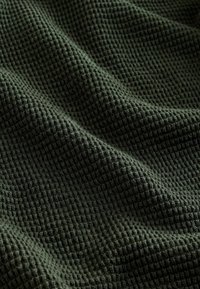 Tejido verde oscuro con un patrón de tejido tipo waffle texturizado, presentado con suaves pliegues y una superficie de rejilla ligeramente elevada.