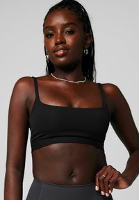 Schwarzer, kurzer Bralette mit dünnen Trägern, glatter Textur und minimalistischem Design. Getragen mit hoch taillierten Hosen, die den Bauch betonen.