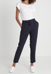 Marinblå byxor med vita prickar, elastisk midja, sidofickor och smalnade ben. Matchas med en enkel vit t-shirt med kort ärm och vita skor.