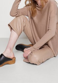 Person sitzt auf dem Boden, trägt einen beigen gerippten Pullover, verkürzte Lederhosen und Clogs mit schwarzem Holzsohle und beigem Rand.