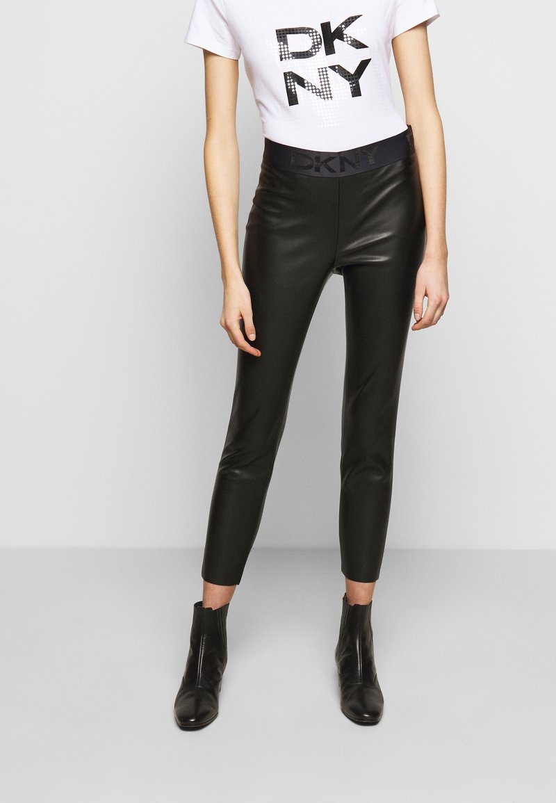 Mannequin portant un pantalon noir en simili cuir taille haute, des bottines noires et un t-shirt blanc ajusté avec le logo "DKNY" en lettres noires.