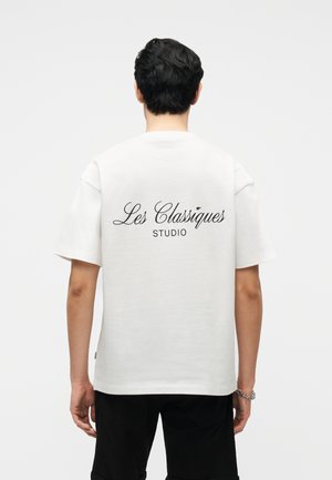 Person med kort, mørkt hår iført en hvid T-shirt med teksten "Les Classiques Studio" på ryggen og sorte bukser, stående mod en ensfarvet baggrund.