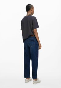 T-shirt noir à manches courtes associé à un jean en denim bleu foncé, présentant des motifs brodés complexes sur la jambe gauche et un patch en cuir à l'arrière.