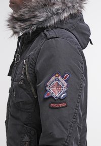 Giacca nera con cappuccio foderato in pelliccia, dotata di molteplici tasche e di un colorato patch ricamato sulla manica che mostra un design geometrico e del testo.