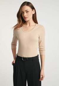 Kobieta z długimi brązowymi włosami, ubrana w beżowy długi rękaw bluzkę włożoną w czarne spodnie z wysokim stanem z czarnym paskiem, stojąca na jednolitym tle.