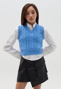 Hellblaues gestricktes Top mit V-Ausschnitt und Zopfmuster, kombiniert mit einem weißen Hemd und einem schwarzen, gewickelten Rock aus Kunstleder.