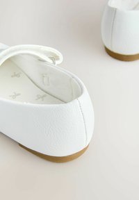 Hvide Mary Jane-flade sko med brune såler på en lys overflade, der viser nærbillede af hæl og ankelstroppens spænde.