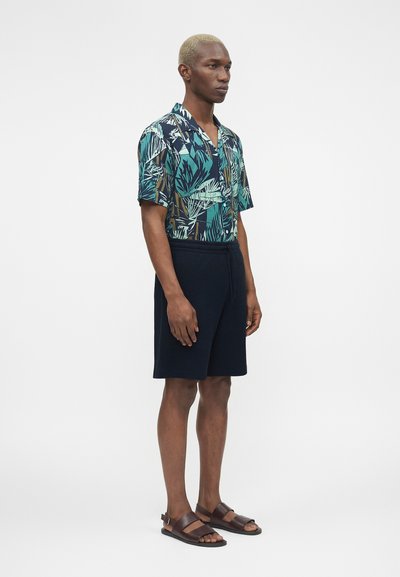 Άνδρας με κοντά ξανθά μαλλιά φοράει κοντομάνικο πουκάμισο με τροπικό σχέδιο, σκούρα σορτς μέχρι το γόνατο και καφέ δερμάτινα σανδάλια, στέκεται στο πλάι.