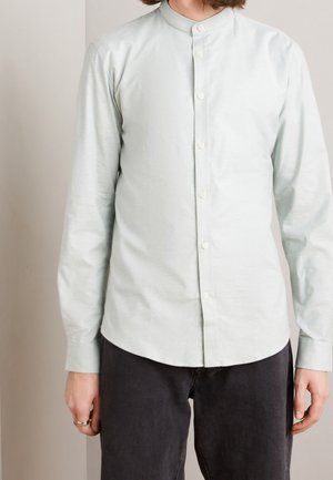 Personne portant une chemise gris clair à col mandarin boutonnée et un pantalon noir, debout devant un fond neutre uni.