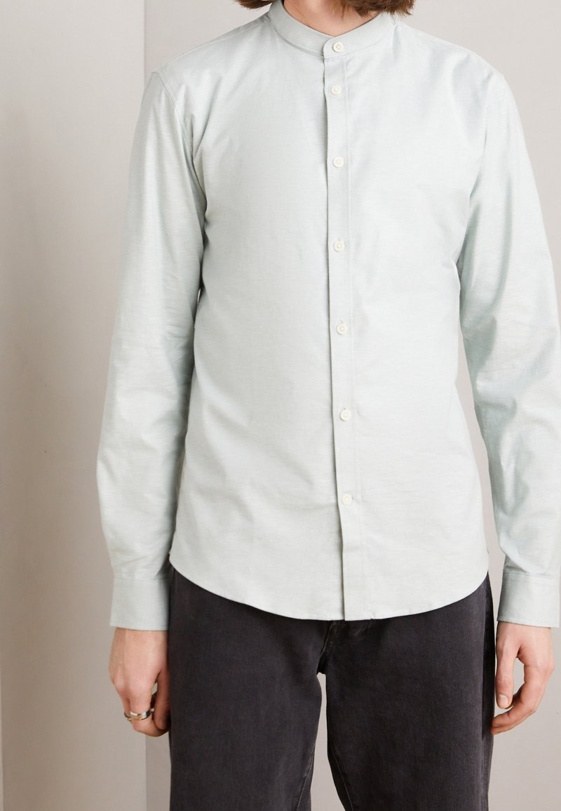 Personne portant une chemise gris clair à col mandarin boutonnée et un pantalon noir, debout devant un fond neutre uni.