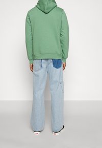 Sudadera verde con capucha combinada con jeans de pierna ancha azul claro que cuentan con un bolsillo de parche. Materiales texturizados, diseño casual y ajuste relajado.