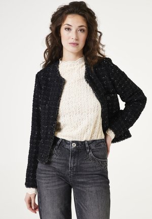 Chaqueta negra texturizada con bordes deshilachados, usada sobre un suéter de punto color crema. Combinada con jeans oscuros que tienen detalles de tachuelas en los bolsillos.