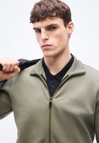 Νέος άνδρας που φοράει ζακέτα με φερμουάρ σε ελιά πράσινο χρώμα με το λογότυπο "BOSS", κρατώντας έναν μαύρο ιμάντα πάνω στον ώμο του, κοιτάζοντας την κάμερα.