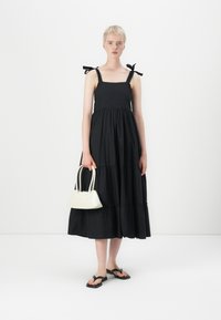 Melnas līdzenošas kleitas ar kvadrātveida izgriezumu un loku siksnām. Modelis tur mazu baltu rokassomu. Valkāts ar melniem flip-flop čībām.