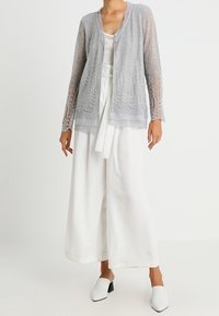 Jasnoszary przezroczysty kardigan z misternymi koronkowymi zdobieniami, zestawiony z białymi spodniami o szerokich nogawkach i białymi butami wsuwanymi.