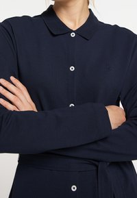 Camicia blu navy con bottoni, maniche lunghe, texture a costine e colletto. Presenta un logo ad ancora e dettagli decorativi con bottoni.