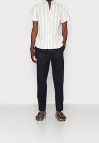 Kortebloemige, lichtbeige en witte gestreepte shirt met een knoopsluiting, gecombineerd met donkerblauwe broeken en olijfgroene sandalen.