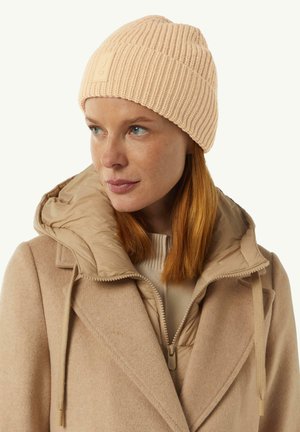 Light pink ribbed knit beanie with a textured finish. Features a small rectangular logo patch. Model wears a beige coat with a hood.