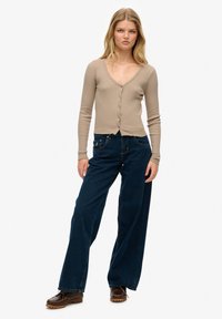 Beżowy, prążkowany kardigan z długim rękawem, z dekoltem w serek i guzikami; noszony z ciemnoniebieskimi, szerokimi spodniami z wysokim stanem i brązowymi butami.