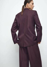 Bordowy, dopasowany garnitur z usztywnioną marynarką z pojedynczym rozcięciem i szerokimi spodniami, wykonany z gładkiej tkaniny z guzikami na rękawach.