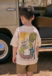 Beige T-Shirt mit kurzen Ärmeln, das bunte, illustrierte Grafiken eines Krokodils und einer Palme zeigt, mit dem Text "Snap Snap Summer" darunter.