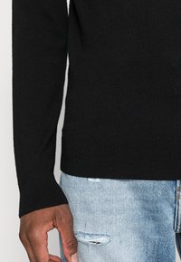 Pull noir en maille avec manches longues, poignets et ourlet côtelés. Associé à un jean déchiré bleu clair, mettant en valeur les coutures détaillées et la texture.