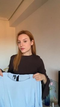 Νεαρή γυναίκα με κόκκινα μαλλιά κρατάει ένα γαλάζιο πουκάμισο εσωτερικά απέναντι σε έναν μπεζ τοίχο με μια μικρή ζωγραφιά με λουλούδια ορατή.