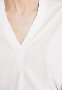 Blouse blanche en coton avec un décolleté en V, présentant une texture lisse et des manches courtes. Le tissu semble léger et respirant.