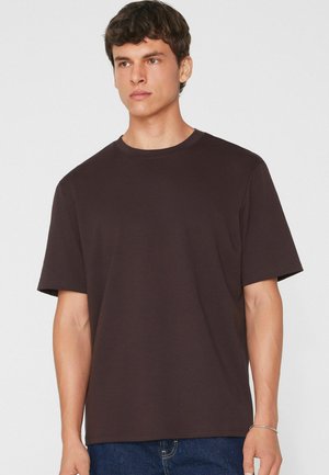 Junger Mann mit kurzen dunklen Haaren, der ein schlichtes dunkelbraunes Kurzarm-T-Shirt und blaue Jeans trägt, steht vor einem hellen Hintergrund.