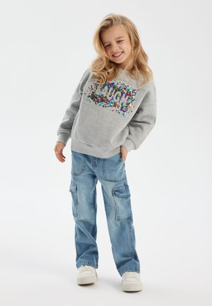 Sudadera gris con un diseño de lentejuelas coloridas que dice "LAUGH", combinada con jeans cargo de color azul claro con bolsillos. El modelo lleva zapatillas blancas.
