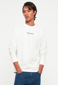 Mann med middels langt mørkt hår hviskler en hvit langermet skjorte med "Calvin Klein" tekst og blå jeans, hender i lommene, nøytral bakgrunn.