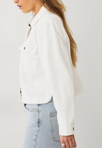 Hvid denimjakke med sidelommer, knaplukning og cropped design. Har en glat tekstur og enkel skrædderarbejde.