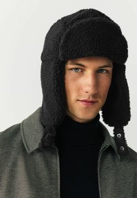 Black fleece trapper hat with ear flaps, textured material, and snap buttons. Worn with a dark turtleneck and patterned collar jacket.