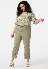 Pantalons vert olive avec taille élastique et poches avant, associés à un blouse à manches courtes et décolleté en V à motifs verts et crème, portés avec des sandales tressées.