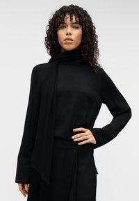 Μαύρο τοπ με μακριά μανίκια, ψηλό γιακά και ρέον ύφασμα, συνδυασμένο με ένα κασκόλ που διαθέτει διακοσμητικές φούντες. Απλός, κομψός σχεδιασμός.