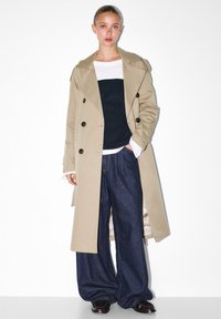 Beżowy płaszcz trench z podwójnymi guzikami, niebieski prążkowany top, szerokie dżinsowe spodnie oraz ciemne buty na białym tle.