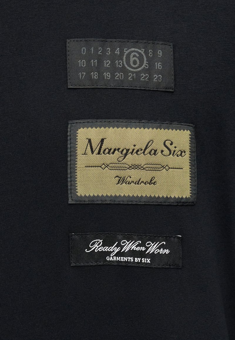 Tissu noir avec trois étiquettes textiles : une étiquette grise avec des chiffres, une étiquette dorée portant l'inscription "Margiela Six" et une étiquette noire "Ready When Worn".