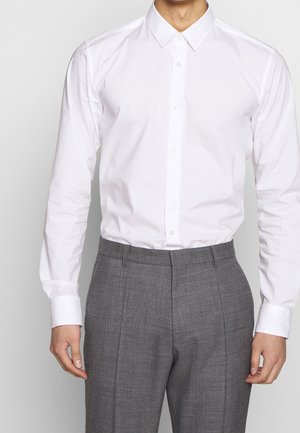 Uomo che indossa una camicia bianca abbottonata infilata in pantaloni eleganti grigi, in piedi davanti a uno sfondo semplice.