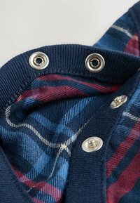 Gros plan sur un tissu à carreaux bleu marine et rouge avec des boutons-pression métalliques sur un bord côtelé, montrant les boutons en position ouverte et fermée.