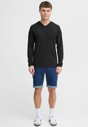 Schwarzes Longsleeve-Shirt mit V-Ausschnitt, kombiniert mit hochgekrempelten Jeansshorts. Das Modell trägt weiße Socken und Sneakers mit schwarz-weißen Akzenten.