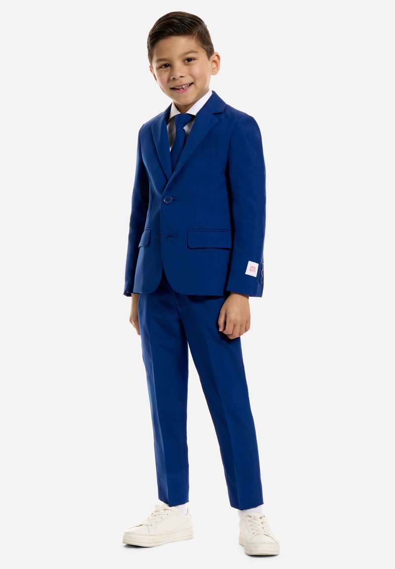 Blauw pak met twee knopen en notch-lapels, gecombineerd met een wit overhemd en een blauwe stropdas. Witte sneakers maken de outfit compleet.