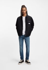 Μαύρο καπιτονέ μπουφάν με φερμουάρ μπροστά, με τσέπη στο στήθος, φορεμένο πάνω από λευκό πουκάμισο, συνδυασμένο με ανοιχτό μπλε τζιν και μαύρα αθλητικά παπούτσια.