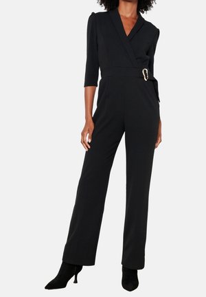 Kobieta ubrana w czarny kombinezon z kopertowym dekoltem, rękawami do połowy przedramienia, boczną klamrą, szerokimi nogawkami oraz czarne kozaki na obcasie z ostrym noskiem.