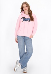 Rózsaszín kapucnis pulcsi tacskó grafikával, húzózsinórokkal és kenguruzsebbel. Világoskék farmerral és ezüst sportcipővel párosítva. Egyszerű háttér.