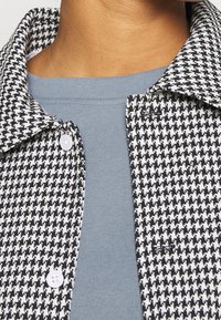 Monokrom houndstooth-skjorta: svart och vit tyg, detalj vid kragen, kombinerad med en mjuk blå rundhalsad topp. Har vita knappar.