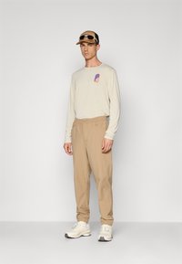 Man standing wearing beige long-sleeve shirt with logo, tan pants with elastic cuffs, white sneakers, and a tan and black cap with sunglasses.