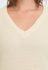 Beige Strickpullover mit V-Ausschnitt, der ein strukturiertes Muster und gerippte Kanten aufweist. Das Material wirkt weich und leicht.