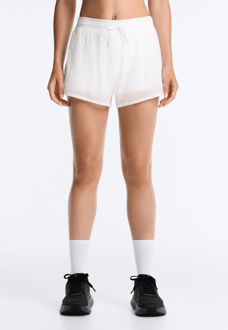 Femme portant un short blanc décontracté, des chaussettes blanches montantes et des chaussures de sport noires, debout devant un fond clair uni.