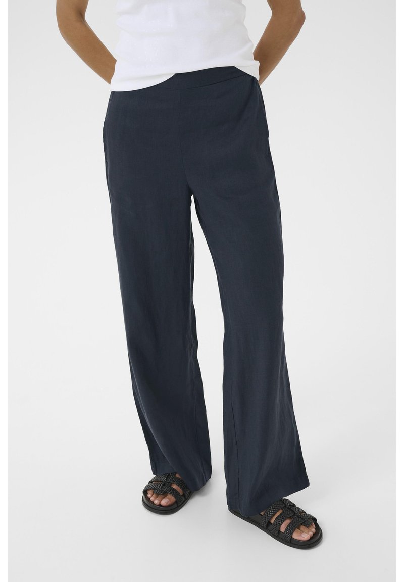 Marineblaue Leinen-Hosen mit weitem Bein verfügen über einen glatten Bund und seitliche Taschen und sind kombiniert mit schwarzen gewebten Sandalen für einen entspannten, lässigen Look.